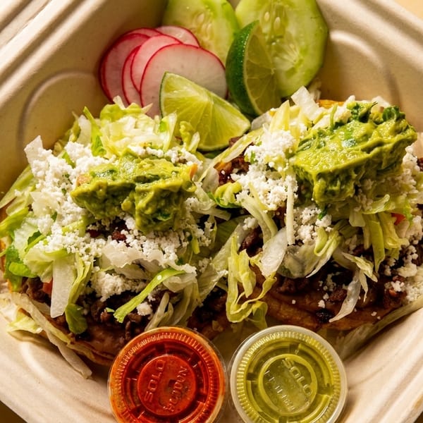 Two sopes topped with meat, lettuce, cheese, and guacamole, with sides of radish, cucumber, and lime.