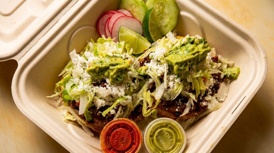 Two sopes topped with meat, lettuce, cheese, and guacamole, with sides of radish, cucumber, and lime.