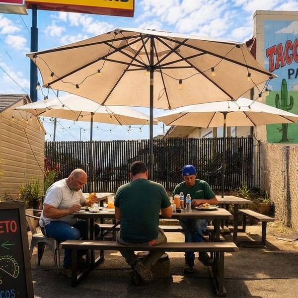 Three men eating at picnic tables under umbrellas on the Taco-Queto Patio, with a taco mural.