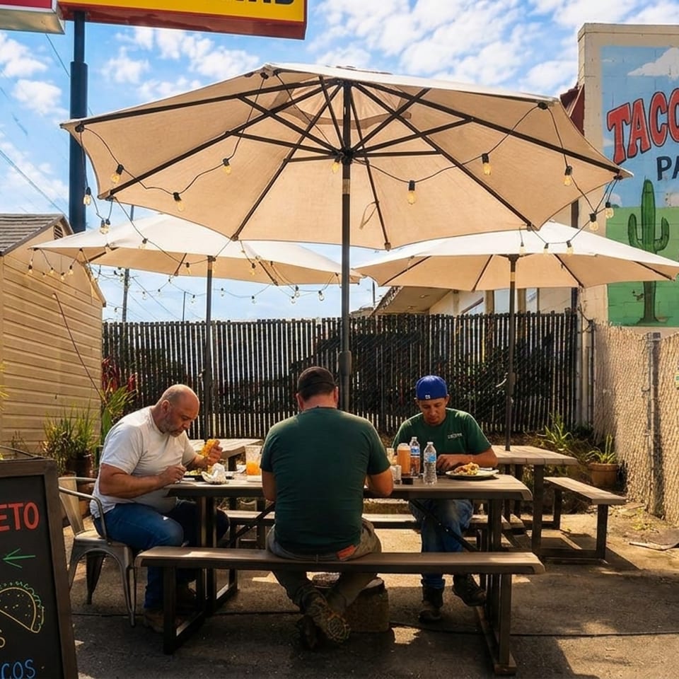 Three men eating at picnic tables under umbrellas on the Taco-Queto Patio, with a taco mural.